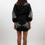 A woman viewed from behind wearing a black satin-like mini dress with white floral embroidery on the shoulders, sleeves, and hem, paired with brown ankle boots, against a light gray studio background.