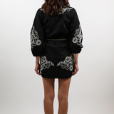 A woman viewed from behind wearing a black satin-like mini dress with white floral embroidery on the shoulders, sleeves, and hem, paired with brown ankle boots, against a light gray studio background.