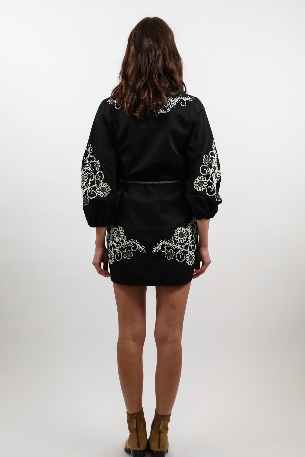 A woman viewed from behind wearing a black satin-like mini dress with white floral embroidery on the shoulders, sleeves, and hem, paired with brown ankle boots, against a light gray studio background.
