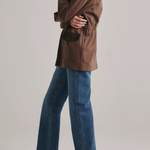 A model shown from the side wearing a brown jacket, blue wide-leg jeans, and black pointed-toe flats against a light gray studio background.