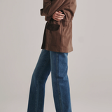 A model shown from the side wearing a brown jacket, blue wide-leg jeans, and black pointed-toe flats against a light gray studio background.