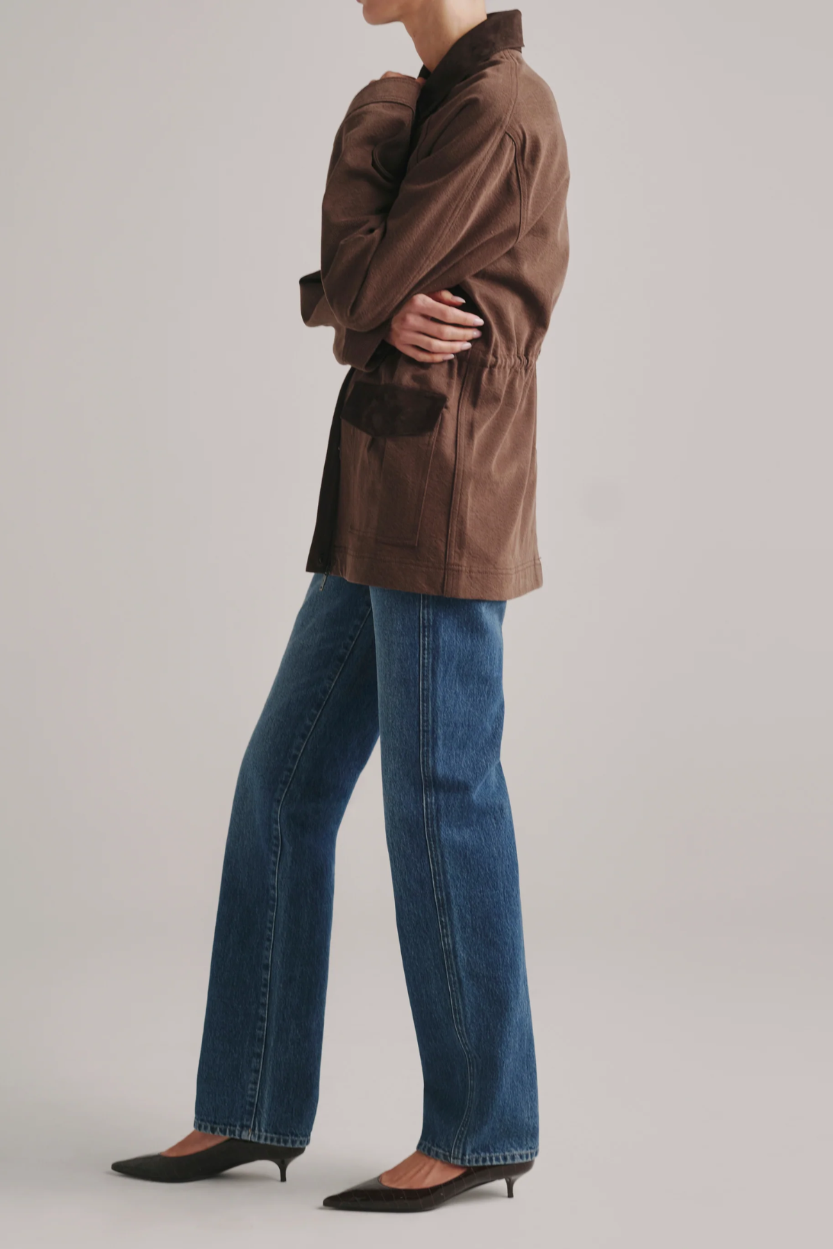 A model shown from the side wearing a brown jacket, blue wide-leg jeans, and black pointed-toe flats against a light gray studio background.