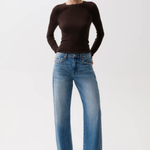 A woman modeling a dark brown long-sleeve knit top and wide-leg blue jeans with decorative silver studs down the sides, paired with black pointed-toe heels, against a plain white studio background.