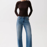 A woman modeling a dark brown long-sleeve knit top and wide-leg blue jeans with decorative silver studs down the sides, paired with black pointed-toe heels, against a plain white studio background.