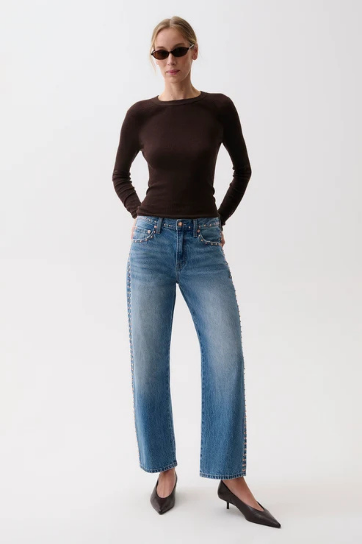 A woman modeling a dark brown long-sleeve knit top and wide-leg blue jeans with decorative silver studs down the sides, paired with black pointed-toe heels, against a plain white studio background.
