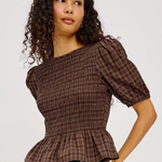A woman wearing a brown smocked plaid top with puff sleeves and a peplum hem, paired with dark blue jeans, against a light gray studio background.