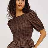 A woman wearing a brown smocked plaid top with puff sleeves and a peplum hem, paired with dark blue jeans, against a light gray studio background.