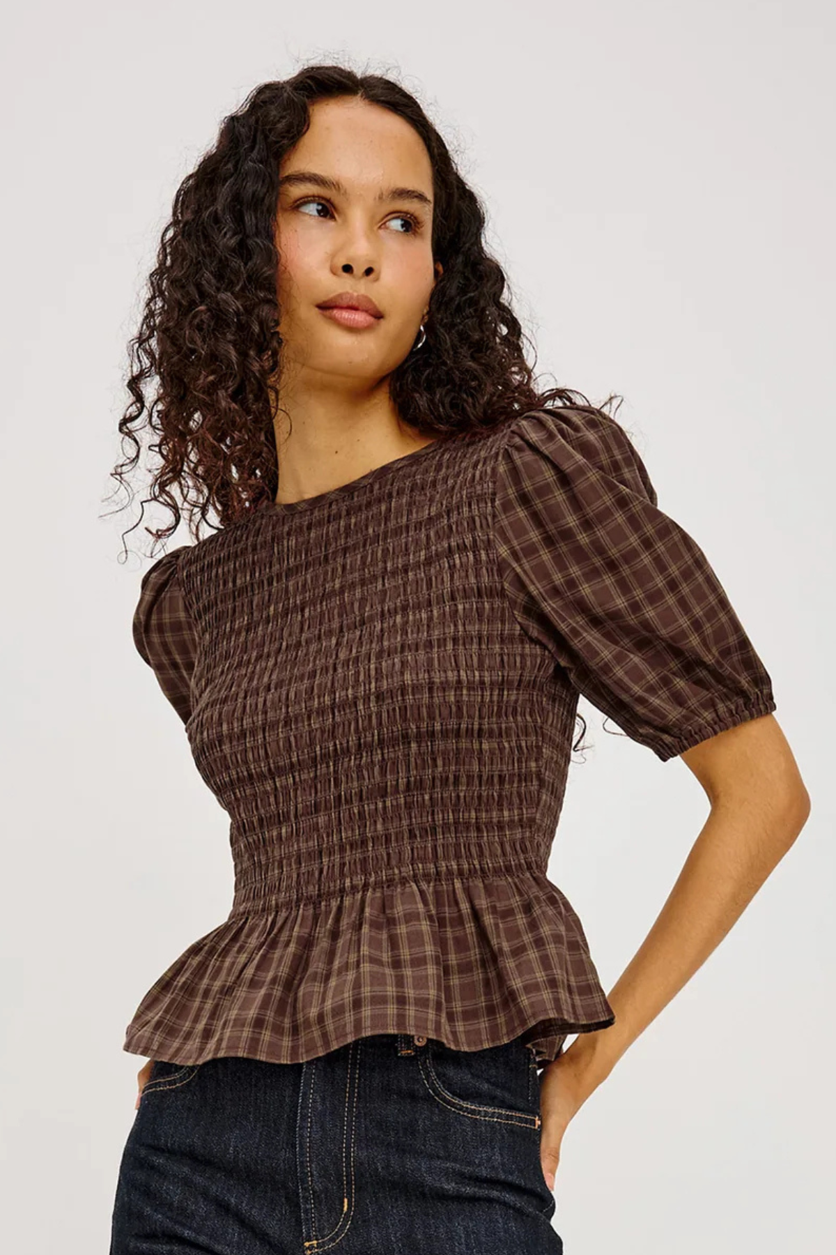 A woman wearing a brown smocked plaid top with puff sleeves and a peplum hem, paired with dark blue jeans, against a light gray studio background.