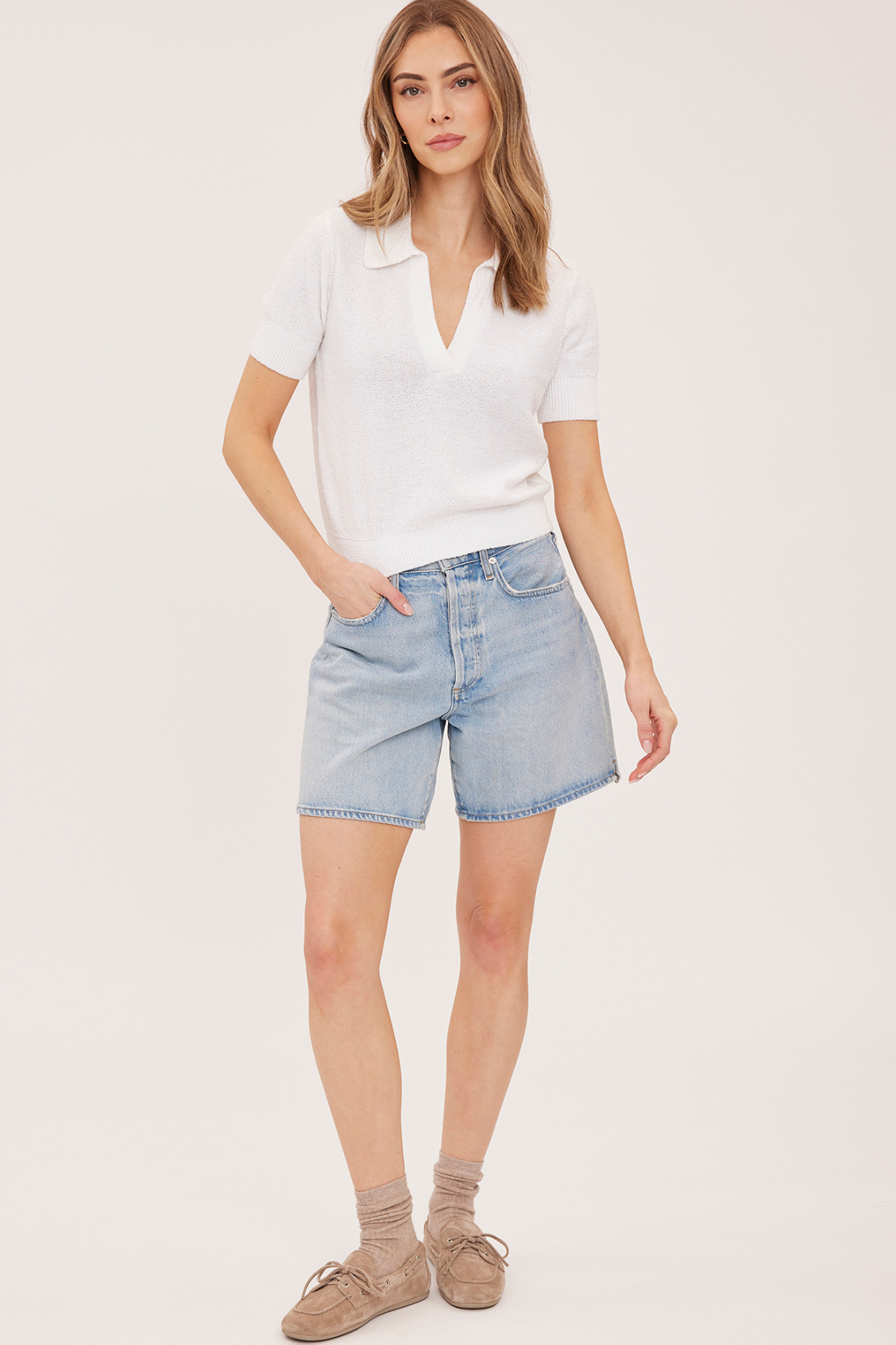 A woman modeling a white short-sleeve polo shirt and light-wash denim shorts against a plain light background.
