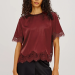 A woman modeling a short-sleeve satin blouse in deep burgundy with lace trim at the sleeves and hem, paired with black satin pants, against a light gray studio background.
