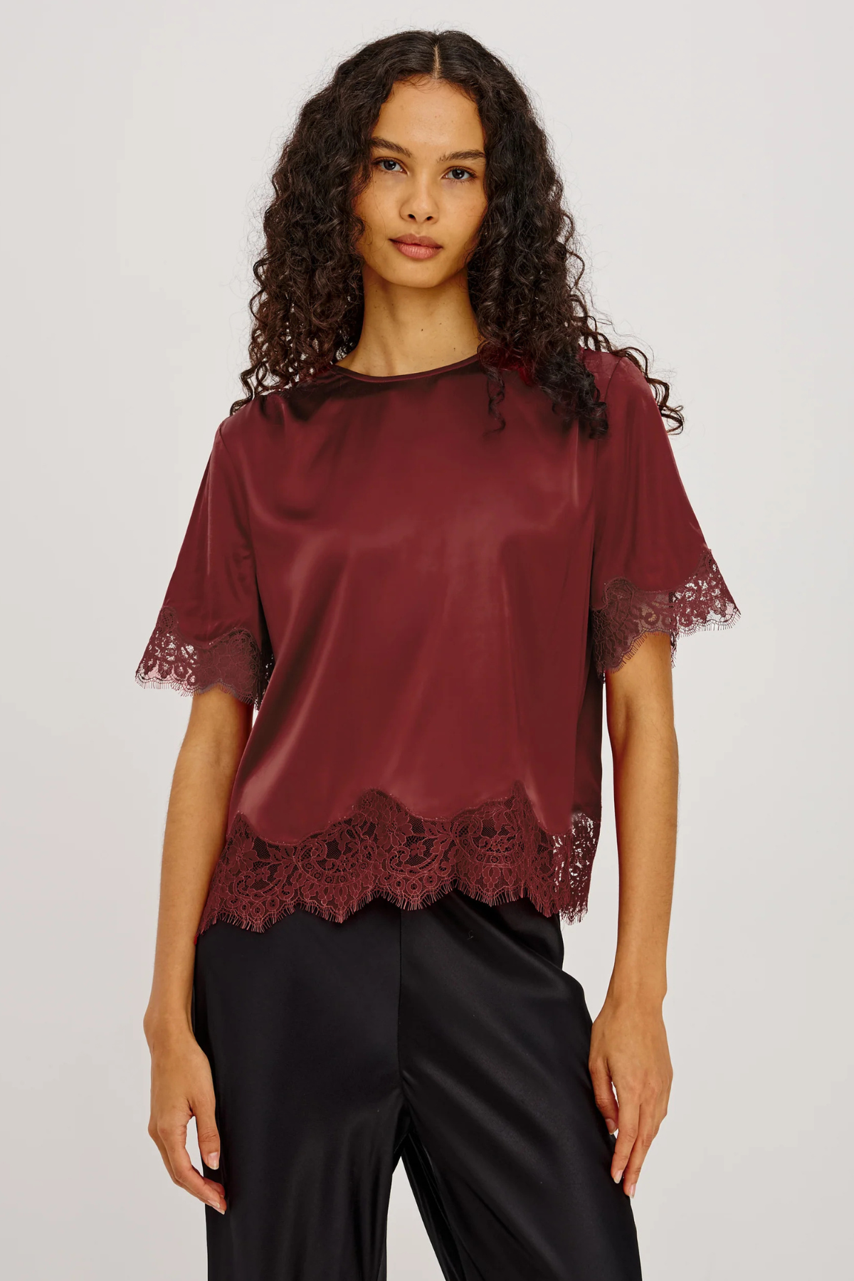 A woman modeling a short-sleeve satin blouse in deep burgundy with lace trim at the sleeves and hem, paired with black satin pants, against a light gray studio background.