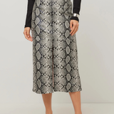 A model wearing a high-waisted midi skirt with a gray and black snakeskin pattern, styled with a black belt, a black ribbed long-sleeve top, and black strappy heels against a light gray studio background.