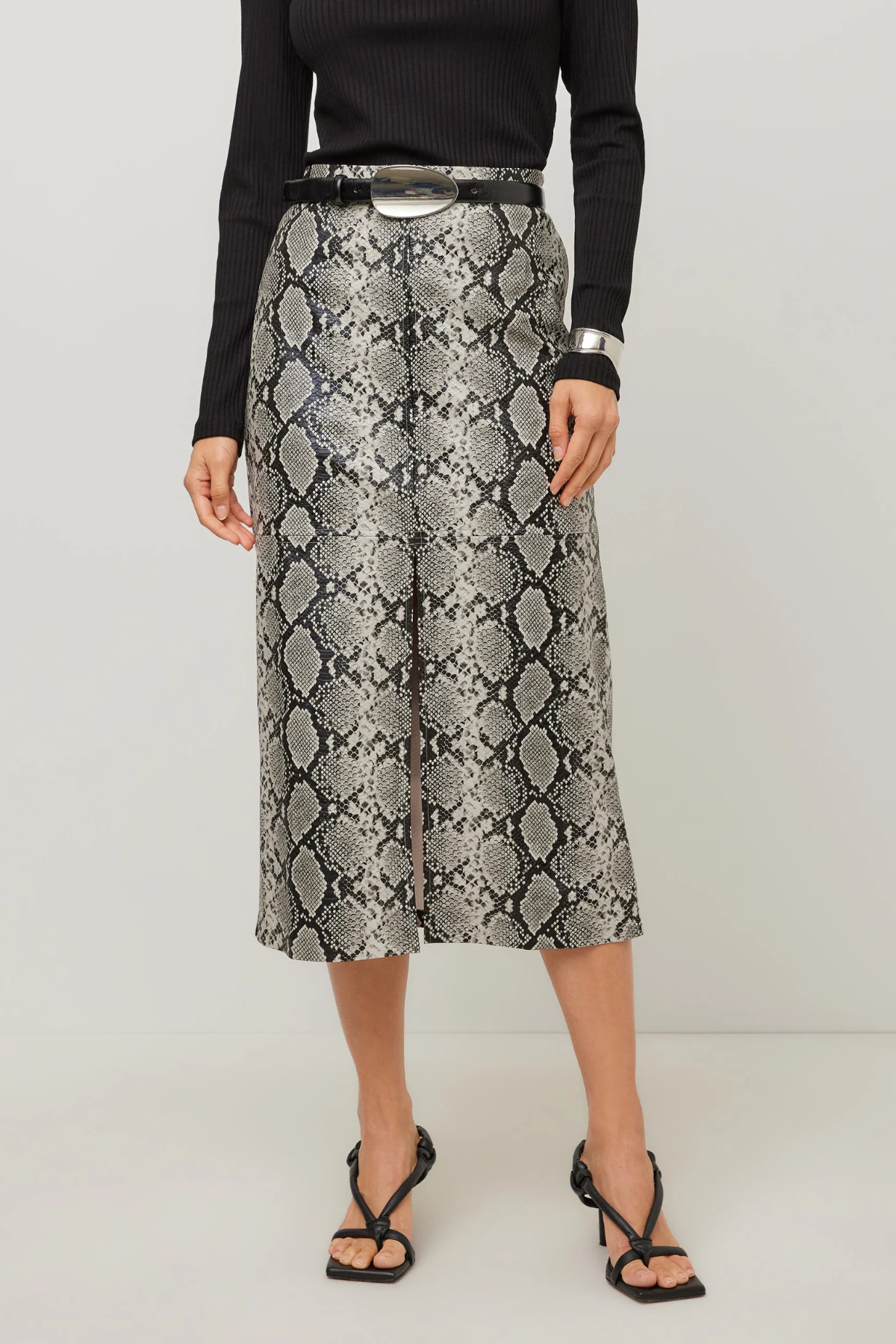 A model wearing a high-waisted midi skirt with a gray and black snakeskin pattern, styled with a black belt, a black ribbed long-sleeve top, and black strappy heels against a light gray studio background.