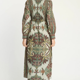 A woman viewed from the back wearing a long-sleeve midi dress with a detailed botanical and floral pattern in olive green, brown, beige, and cream, paired with beige high-heeled sandals against a plain white studio background.