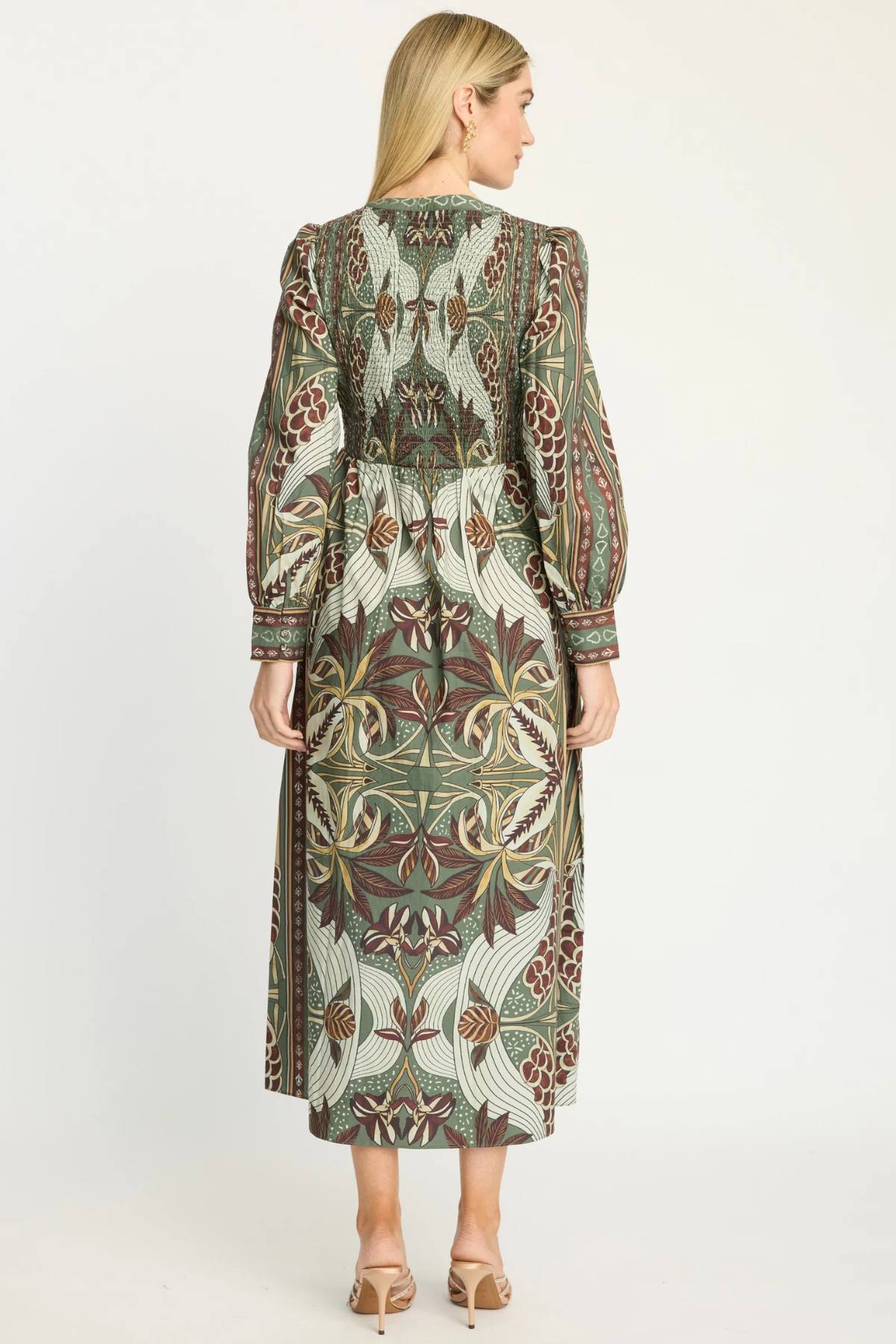 A woman viewed from the back wearing a long-sleeve midi dress with a detailed botanical and floral pattern in olive green, brown, beige, and cream, paired with beige high-heeled sandals against a plain white studio background.