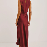 A woman modeling a sleeveless, floor-length burgundy satin dress from the back, featuring a small keyhole cutout at the upper back, set against a plain white studio background.