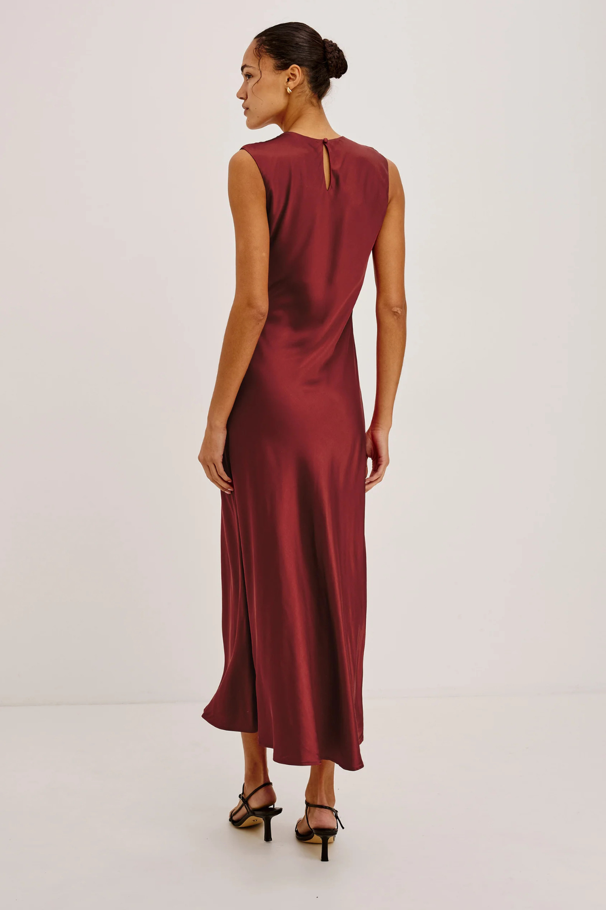A woman modeling a sleeveless, floor-length burgundy satin dress from the back, featuring a small keyhole cutout at the upper back, set against a plain white studio background.