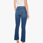 Rear view of a model wearing blue flared jeans, a cream short-sleeve top, and metallic high heels against a light gray studio background.