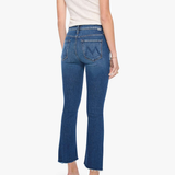 Rear view of a model wearing blue flared jeans, a cream short-sleeve top, and metallic high heels against a light gray studio background.