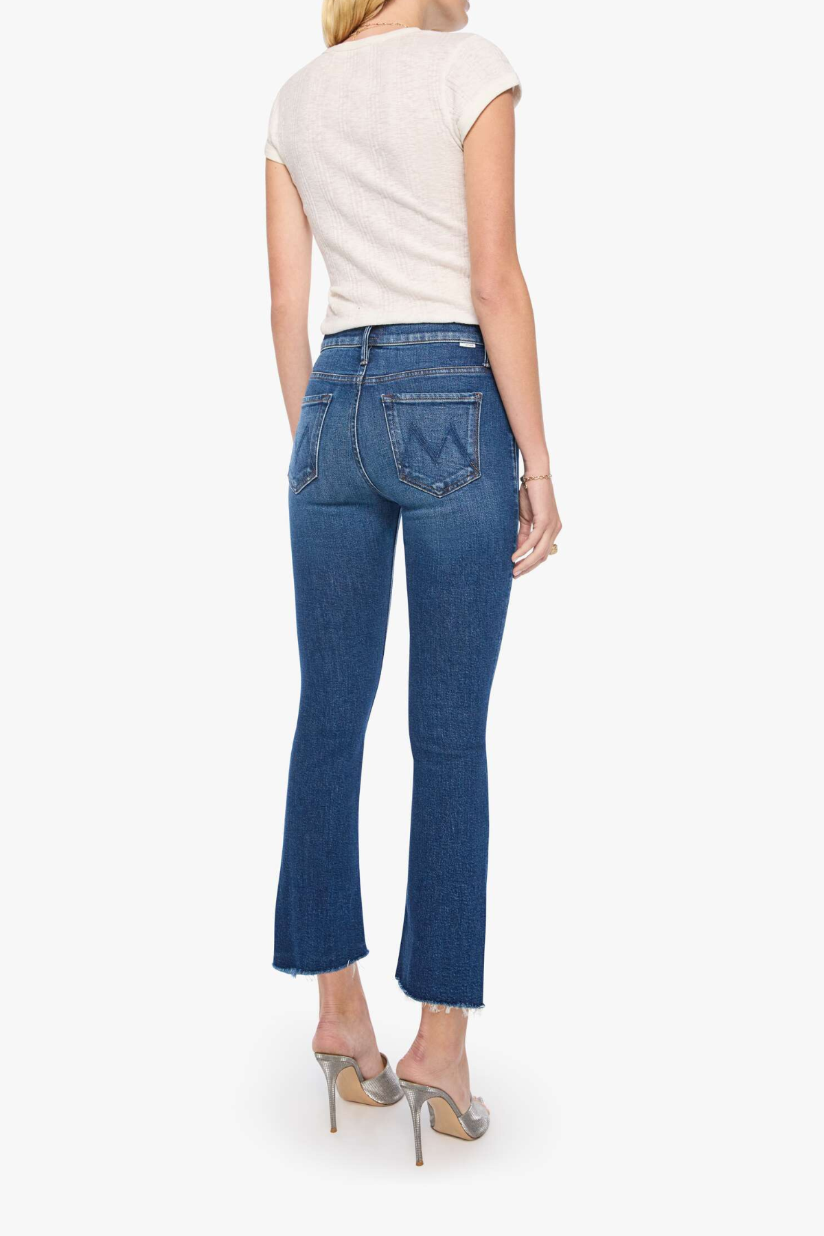 Rear view of a model wearing blue flared jeans, a cream short-sleeve top, and metallic high heels against a light gray studio background.