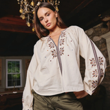 A woman wearing a cream-colored long-sleeve blouse with intricate floral embroidery on the chest and sleeves, paired with olive green cargo pants, standing in a rustic room with a chandelier overhead.