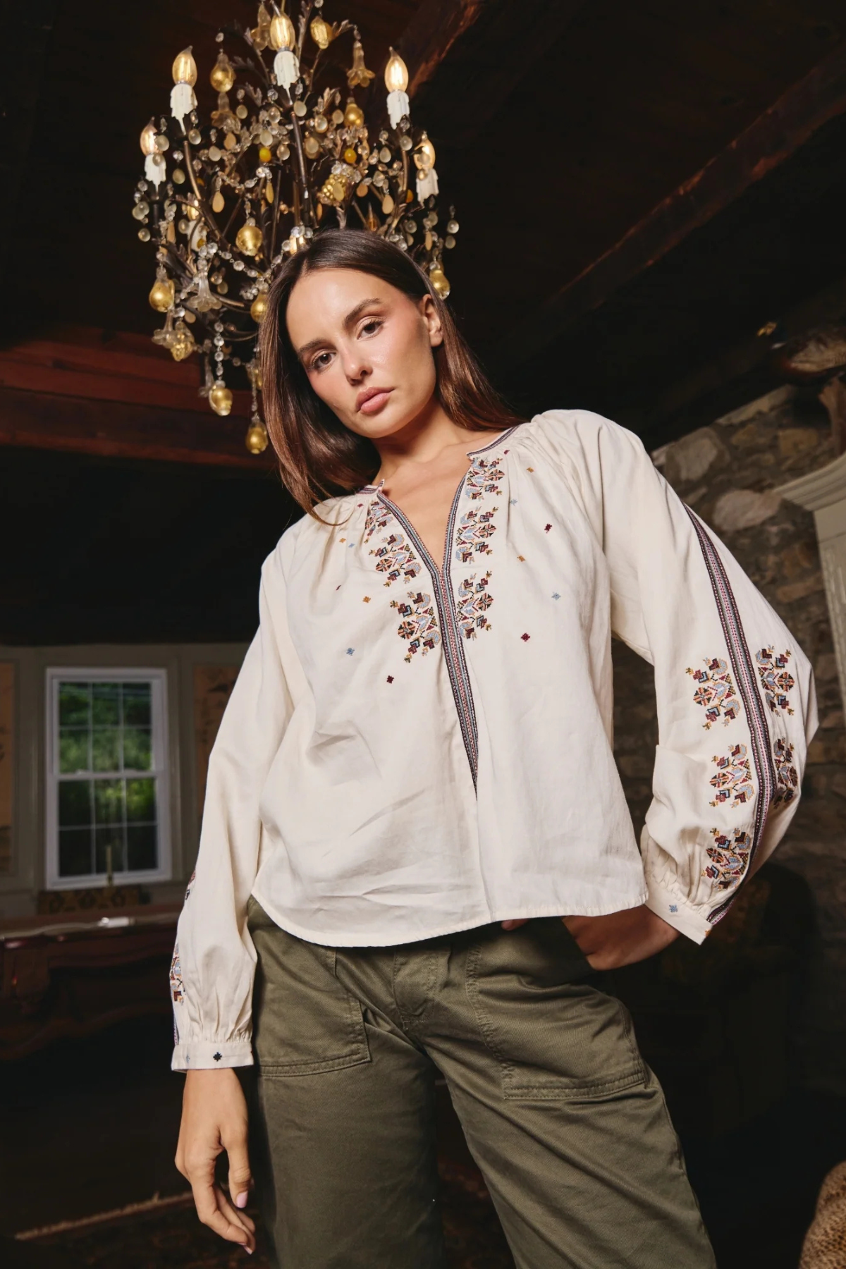 A woman wearing a cream-colored long-sleeve blouse with intricate floral embroidery on the chest and sleeves, paired with olive green cargo pants, standing in a rustic room with a chandelier overhead.