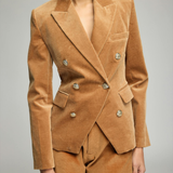 A woman wearing a camel brown velvet double-breasted blazer and matching shorts against a neutral gray studio background.