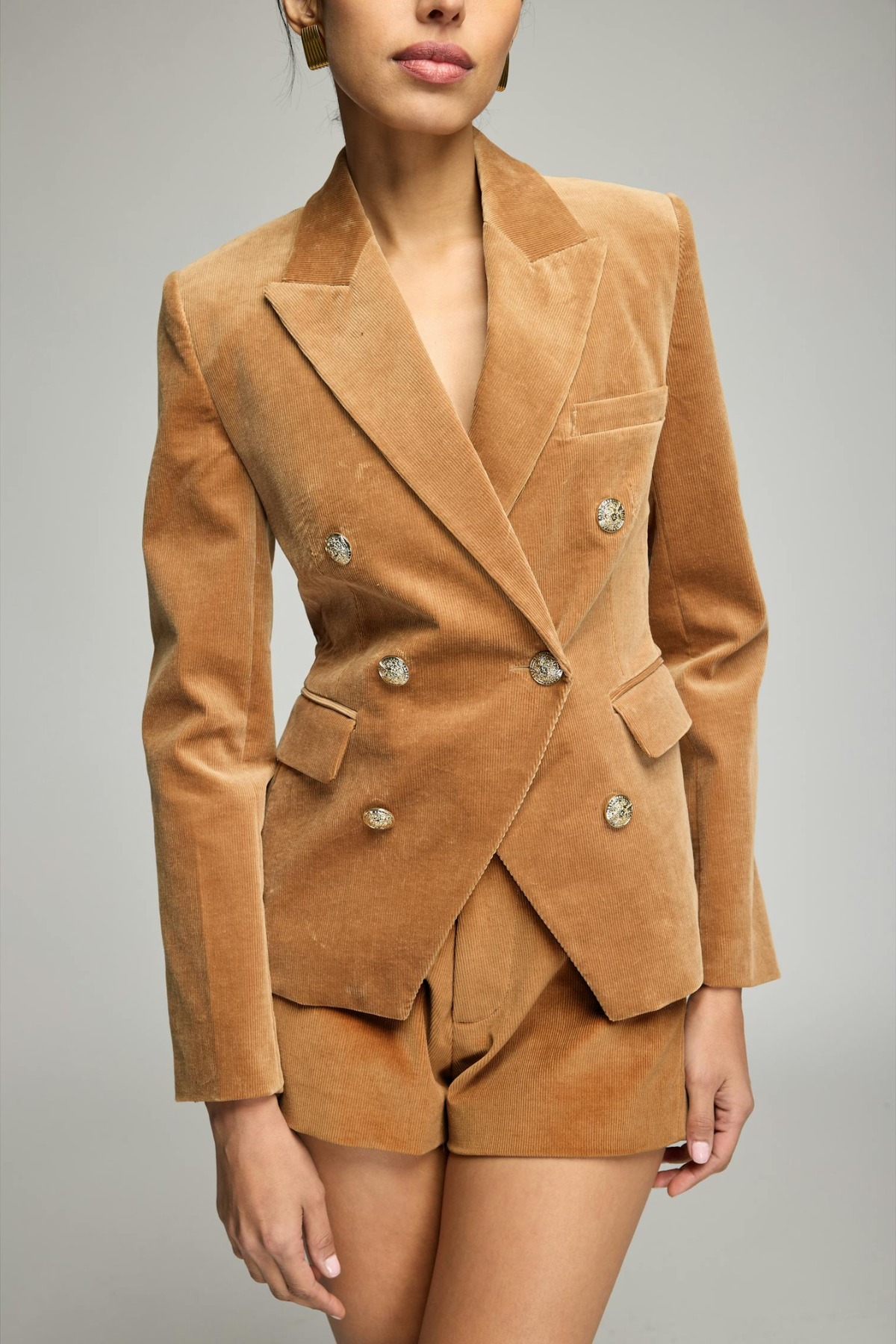 A woman wearing a camel brown velvet double-breasted blazer and matching shorts against a neutral gray studio background.