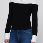 A woman wearing a black off-the-shoulder knit sweater with contrasting white collar and cuffs, paired with blue jeans, against a light gray studio background.