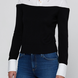 A woman wearing a black off-the-shoulder knit sweater with contrasting white collar and cuffs, paired with blue jeans, against a light gray studio background.
