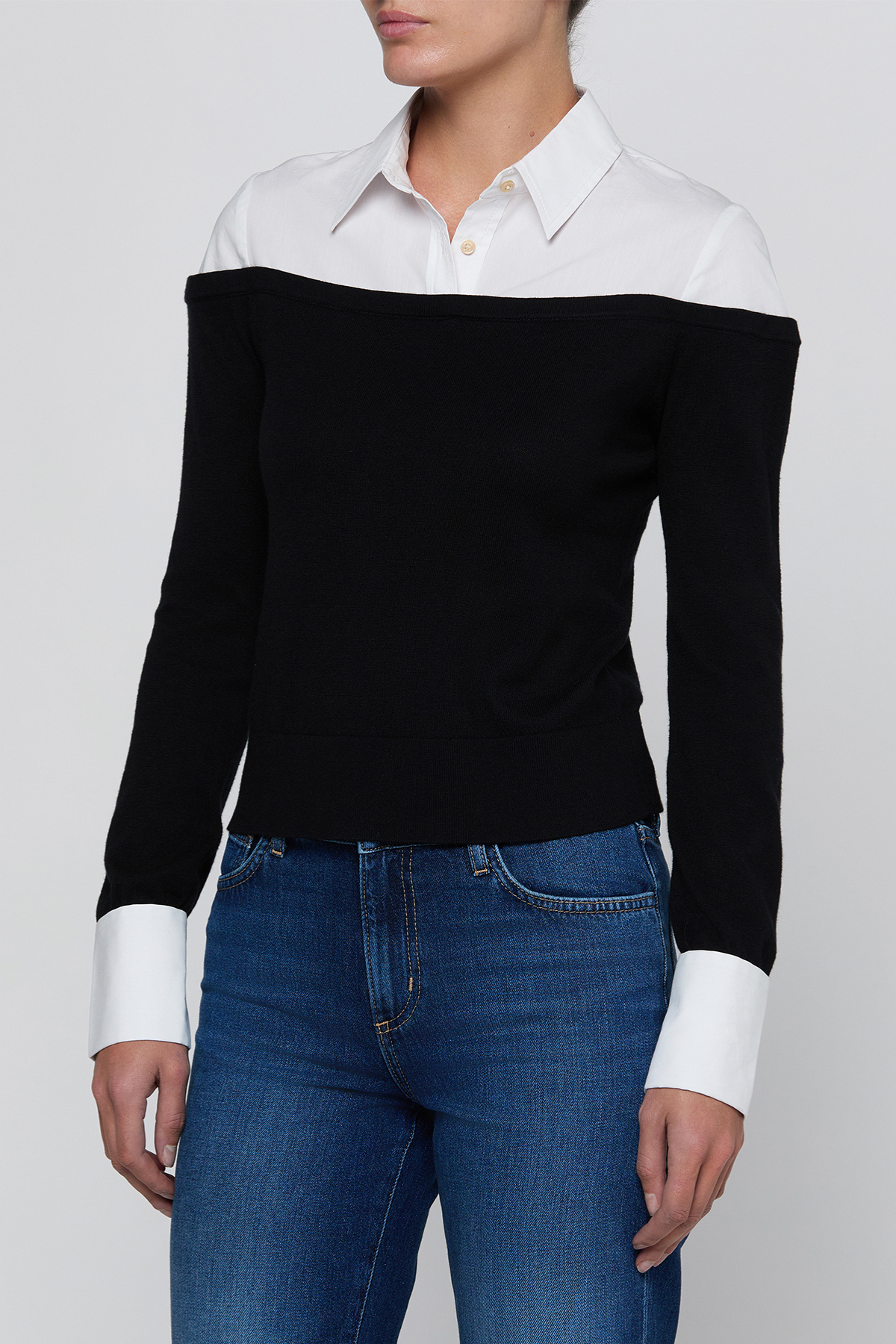 A woman wearing a black off-the-shoulder knit sweater with contrasting white collar and cuffs, paired with blue jeans, against a light gray studio background.