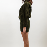 A woman shown in profile wearing a dark olive green velvet long-sleeve top and matching shorts against a plain light gray studio background.