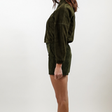A woman shown in profile wearing a dark olive green velvet long-sleeve top and matching shorts against a plain light gray studio background.