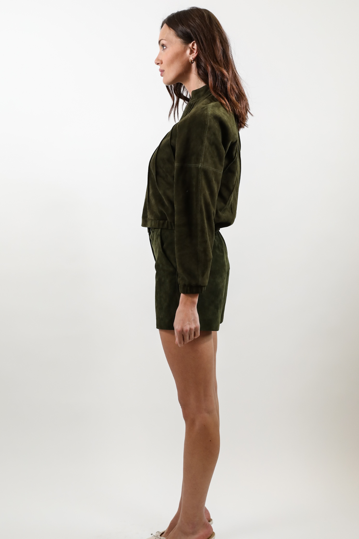 A woman shown in profile wearing a dark olive green velvet long-sleeve top and matching shorts against a plain light gray studio background.