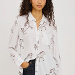 A woman with curly dark hair wearing a white button-up shirt with a brown horse print, paired with dark blue jeans, against a plain light gray background.
