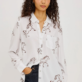 A woman with curly dark hair wearing a white button-up shirt with a brown horse print, paired with dark blue jeans, against a plain light gray background.