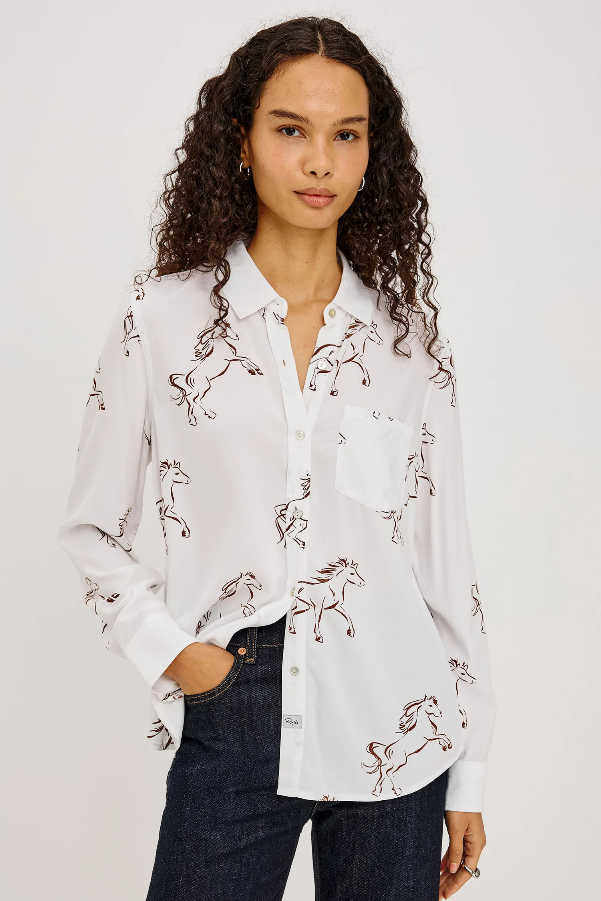 A woman with curly dark hair wearing a white button-up shirt with a brown horse print, paired with dark blue jeans, against a plain light gray background.