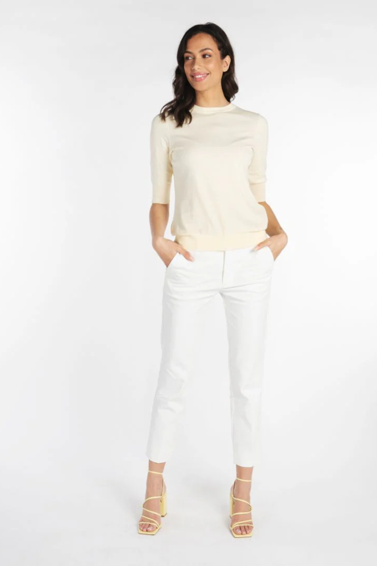 A woman modeling a cream-colored short-sleeve knit top and white cropped trousers with beige strappy heels, standing against a plain white studio background.