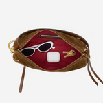 An open brown leather pouch or clutch with a deep red interior, shown on a light gray background. Inside, white sunglasses, a white earbud case, and two keys are arranged as props.