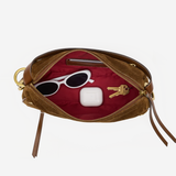 An open brown leather pouch or clutch with a deep red interior, shown on a light gray background. Inside, white sunglasses, a white earbud case, and two keys are arranged as props.