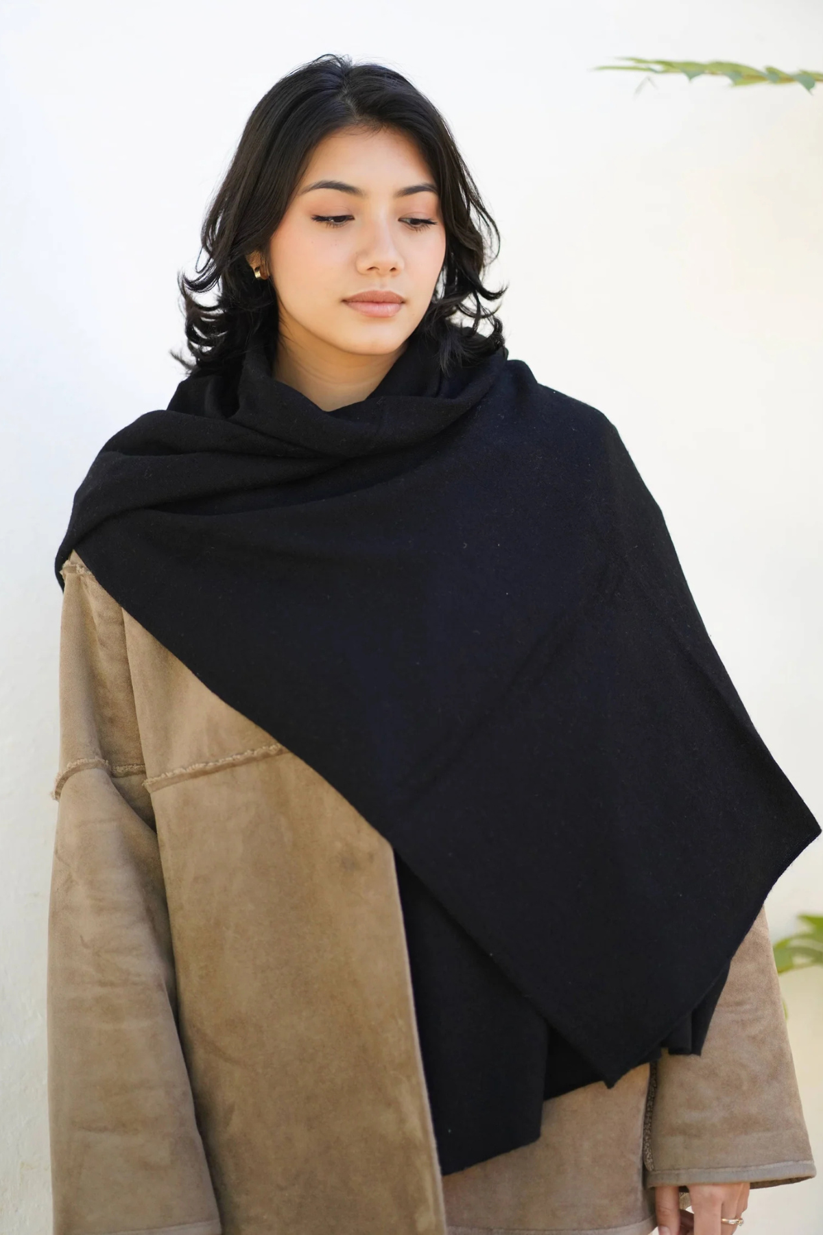 A woman wearing a large black shawl draped over her shoulders, layered over a beige suede jacket, against a clean white background.