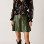 Woman wearing a floral blouse, green leather skirt, and black boots on a plain background