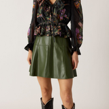 Woman wearing a floral blouse, green leather skirt, and black boots on a plain background