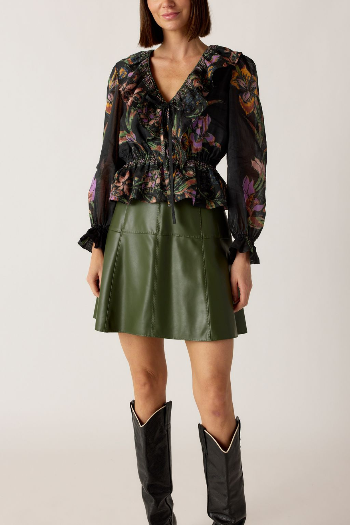 Woman wearing a floral blouse, green leather skirt, and black boots on a plain background