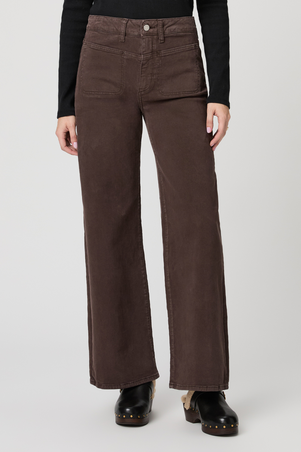A model wearing a pair of dark brown wide-leg corduroy pants, styled with a black long-sleeve top and black leather clogs, against a light gray studio background.
