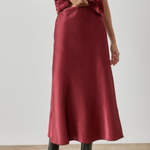 A model wearing a satin midi skirt in a deep burgundy red, paired with a matching short-sleeve top and black ankle boots, standing against a neutral studio background.