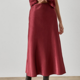 A model wearing a satin midi skirt in a deep burgundy red, paired with a matching short-sleeve top and black ankle boots, standing against a neutral studio background.