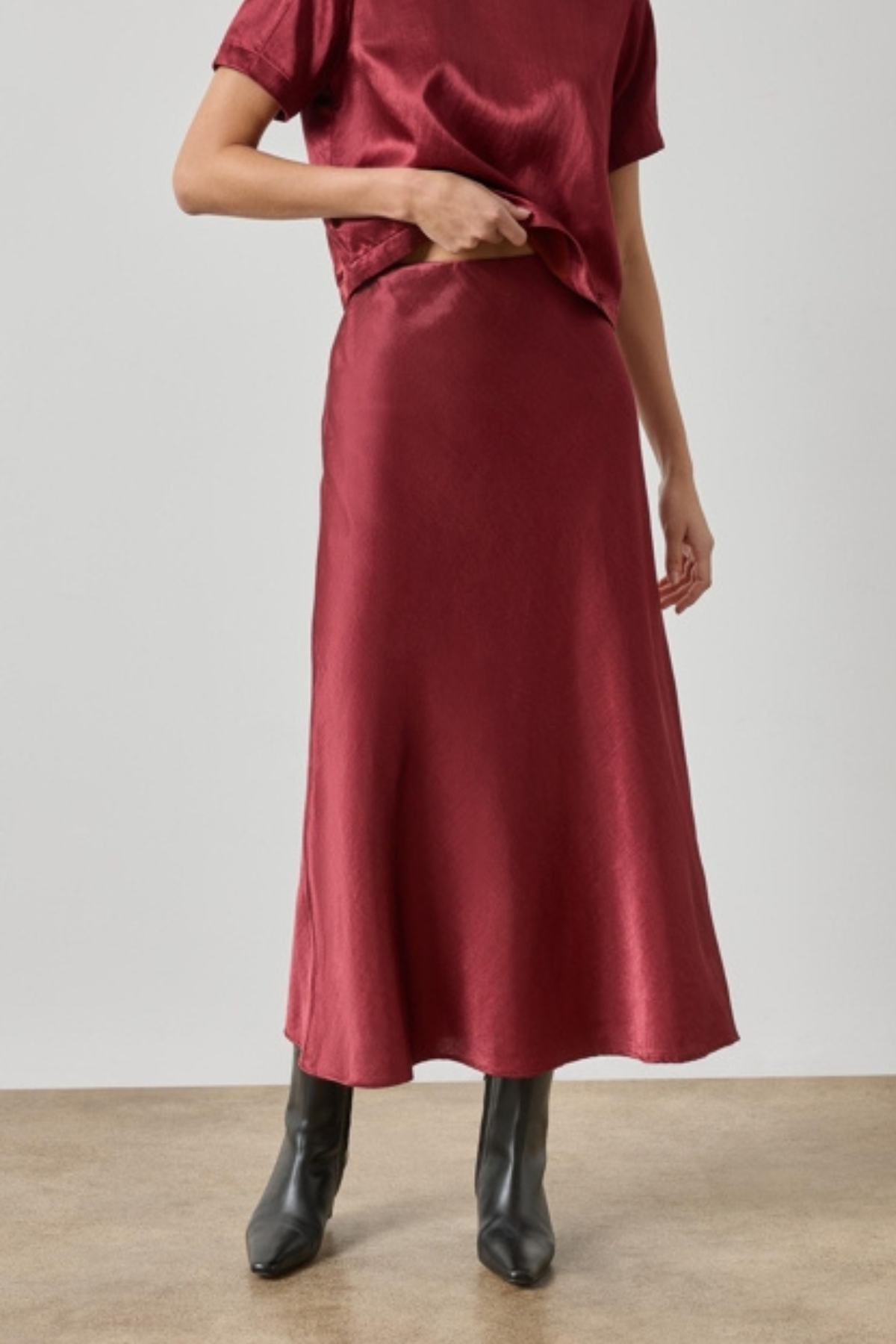 A model wearing a satin midi skirt in a deep burgundy red, paired with a matching short-sleeve top and black ankle boots, standing against a neutral studio background.