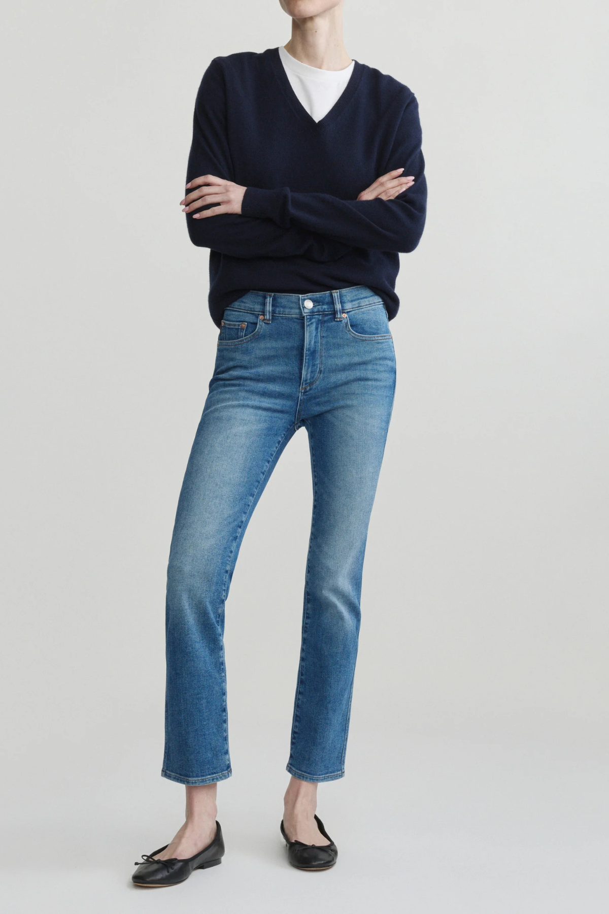 A model wearing a navy blue v-neck sweater, a white undershirt, blue cropped jeans, and black flats, standing with arms crossed against a light gray studio background.