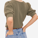 A woman wearing a ribbed short-sleeve knit top in olive green, paired with light blue jeans, shown from a side-back angle against a plain light background.